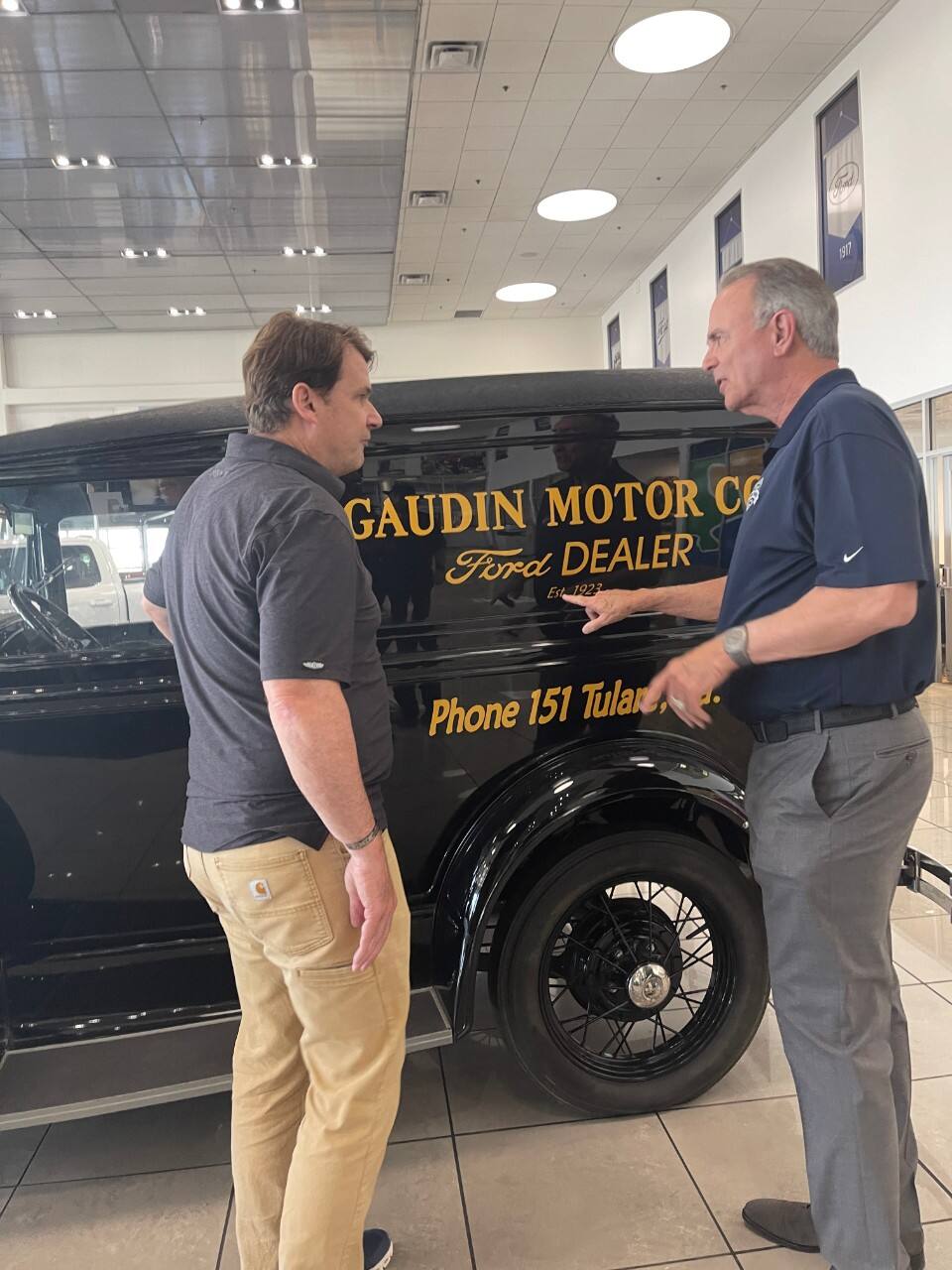 Two men discuss a vintage black Ford vehicle in a dealership showroom.